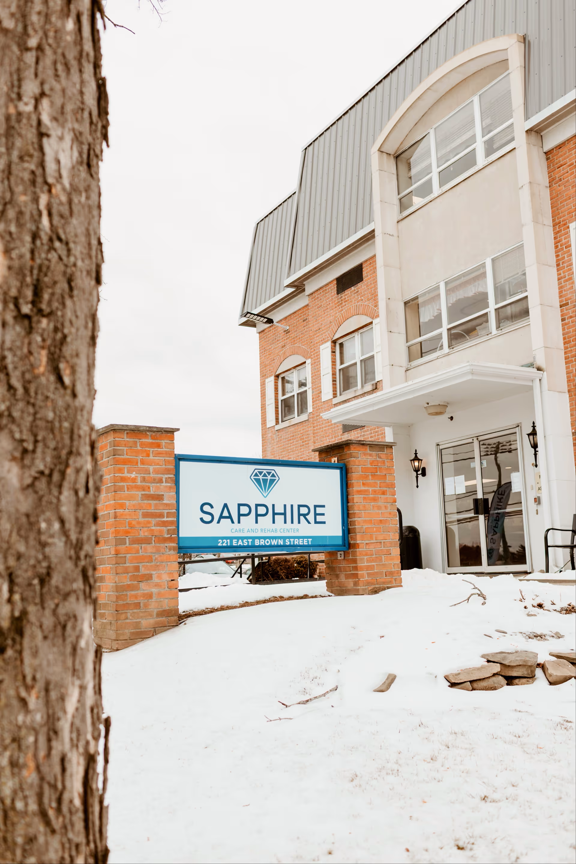 Snow covers a brick senior living building with a glass entry and Sapphire sign.