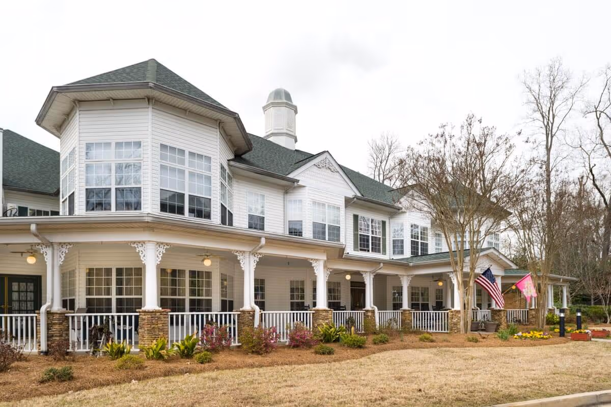 A large white senior living facility with a wraparound porch, lanterns, and flags.