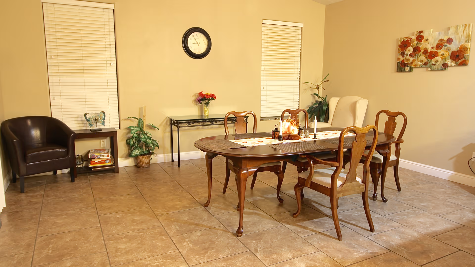 A dining room with a wooden dining table and six chairs, two windows with blinds, a wall clock, a small table with a vase of flowers, a brown leather chair, a potted plant, and a floral painting on the wall.