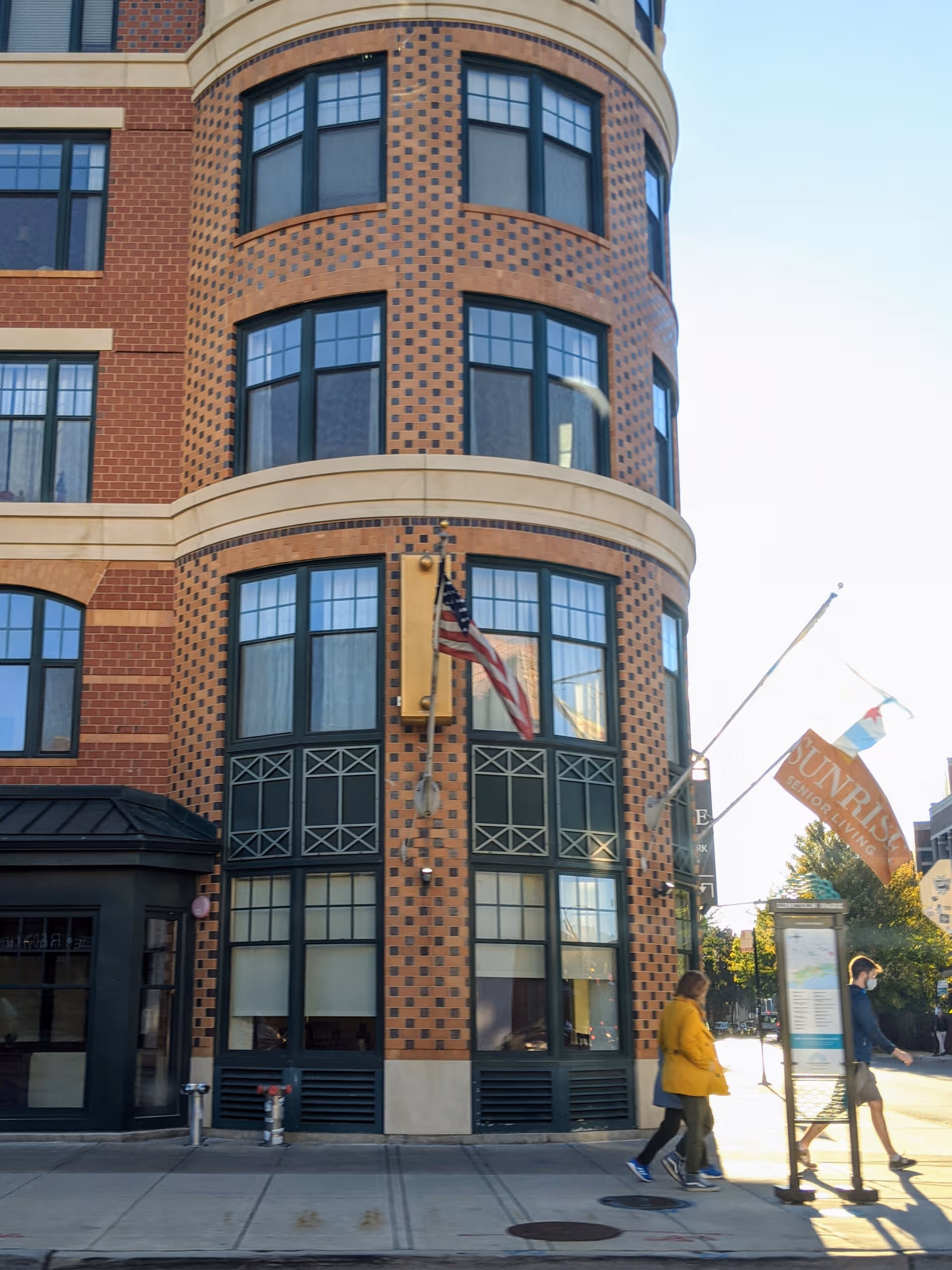 Brick building with curved corner, large windows, flag, and Sunrise sign on sidewalk.