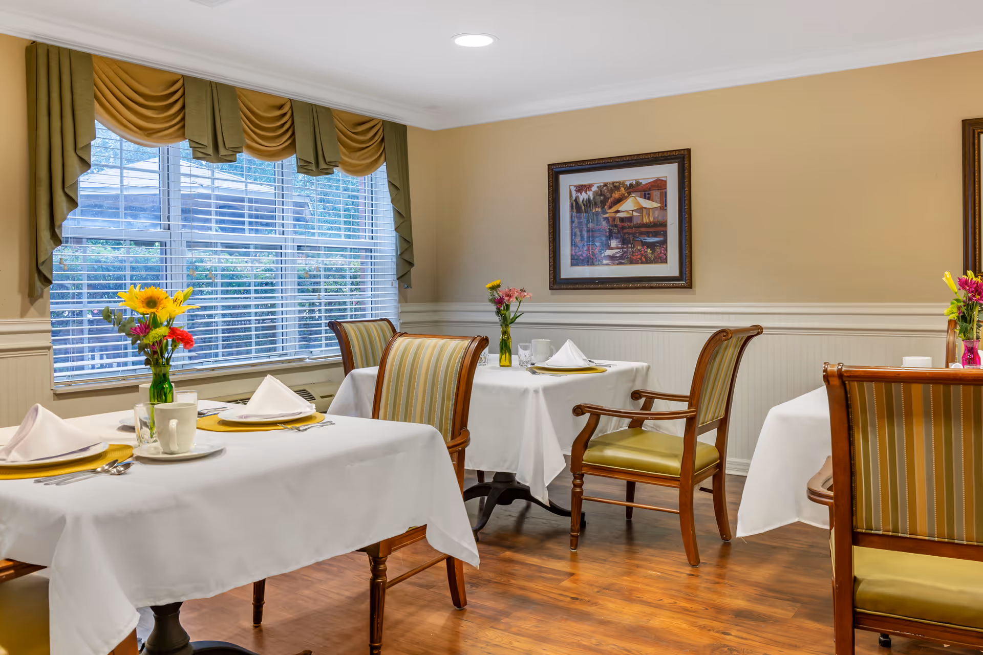 Bright dining room with white-tablecloth tables, floral centerpieces, and striped upholstered chairs.