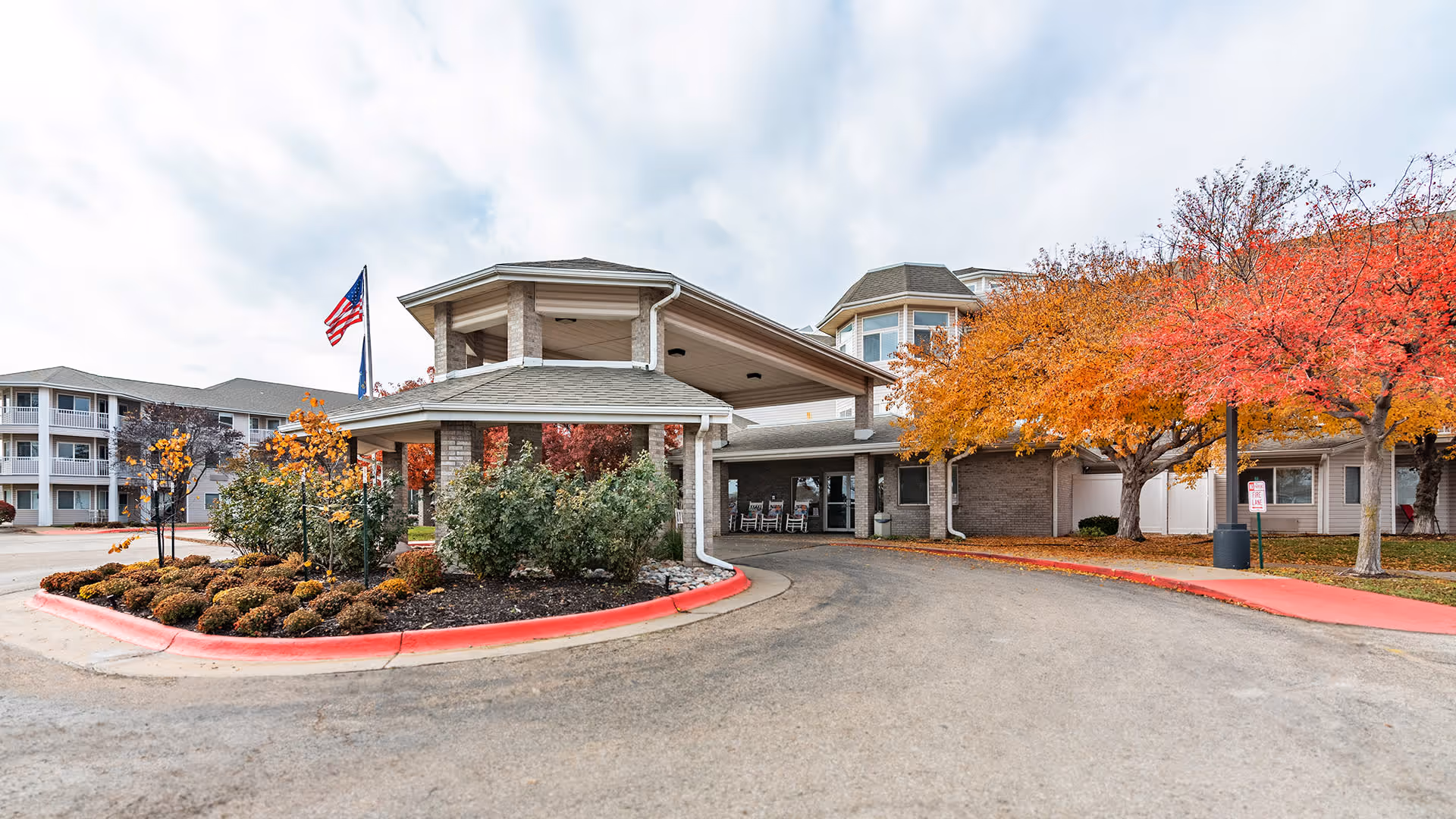 Exterior view of Holiday Thornton Place senior living facility entrance with a covered driveway, landscaped bushes, and trees with autumn foliage. An American flag is visible on a flagpole near the entrance.