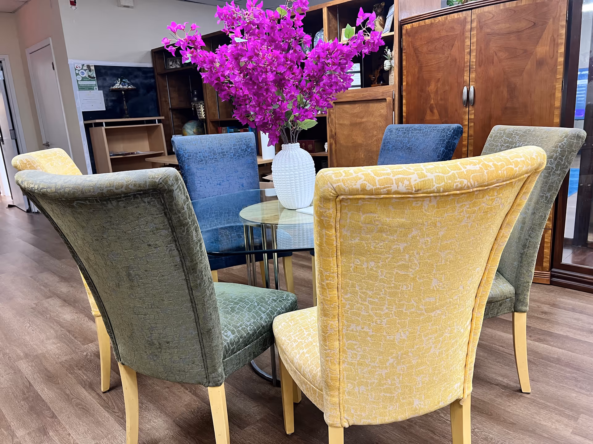 Colorful upholstered chairs surround a glass table with a white vase and purple flowers.