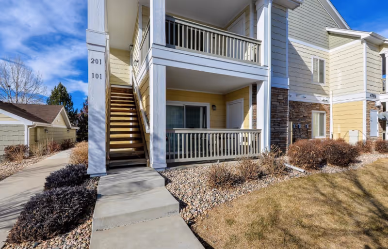 Exterior view of a two-story apartment building showing an exterior staircase, covered ground-floor patio, upper balcony, and landscaped walkway.