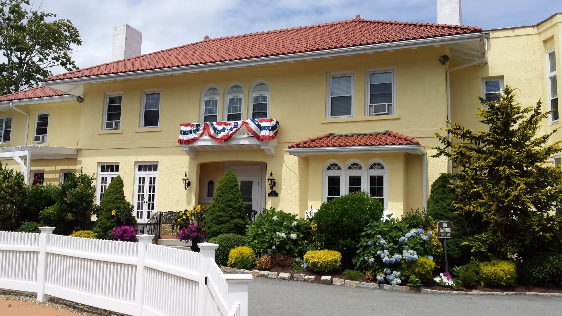 Yellow two-story nursing home front with red-tiled roof, bunting, and manicured garden.