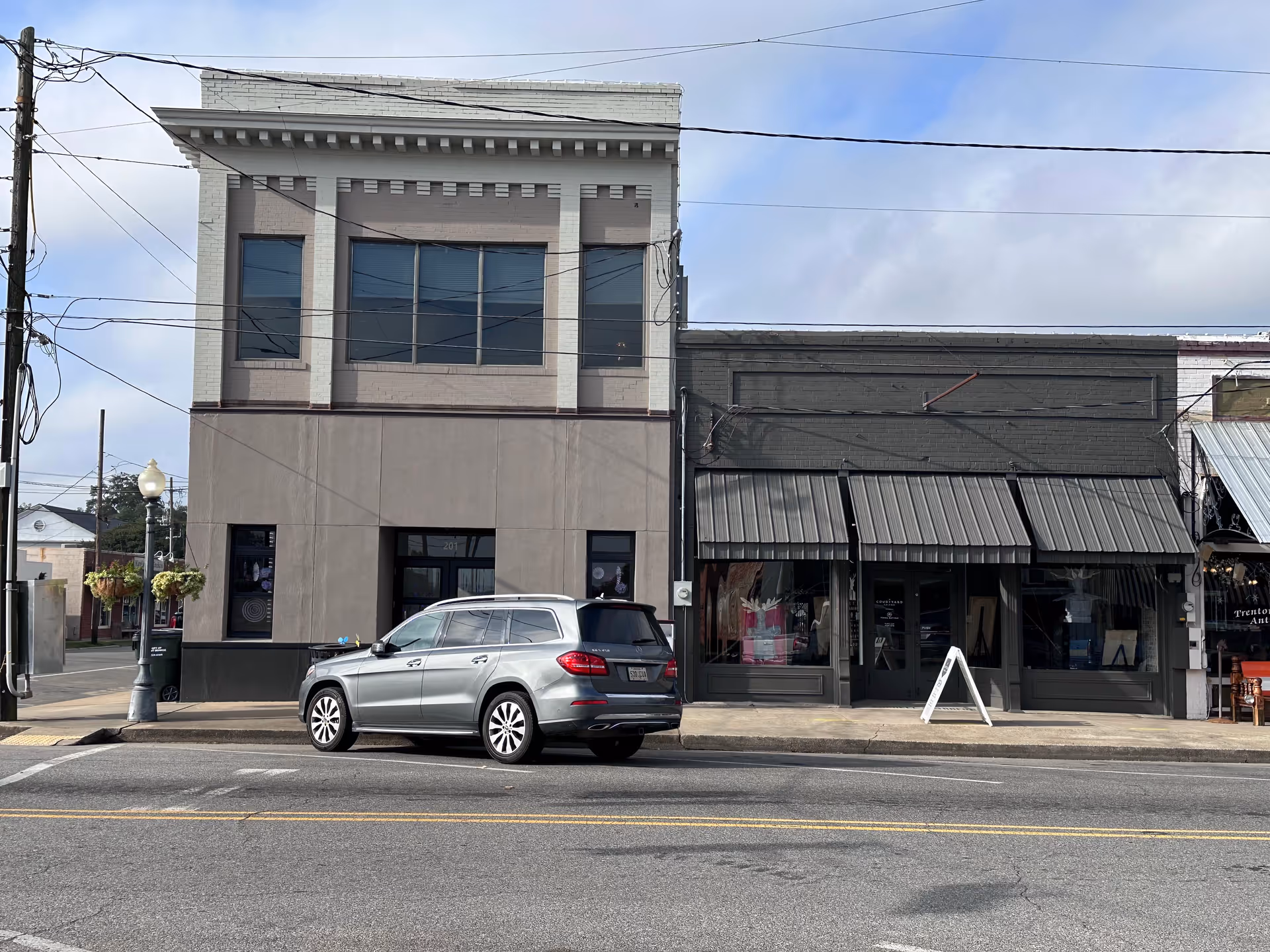 Street view of storefronts with a silver SUV parked along the curb.