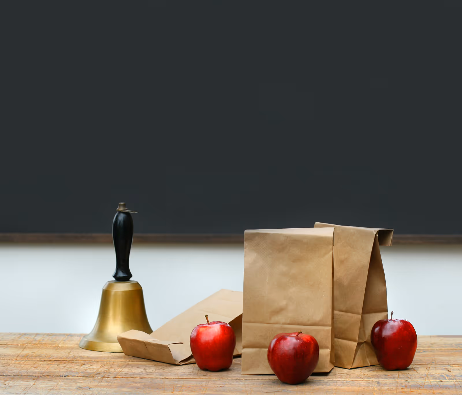 A wooden table with a bell, brown paper bags, and red apples in front of a dark chalkboard.