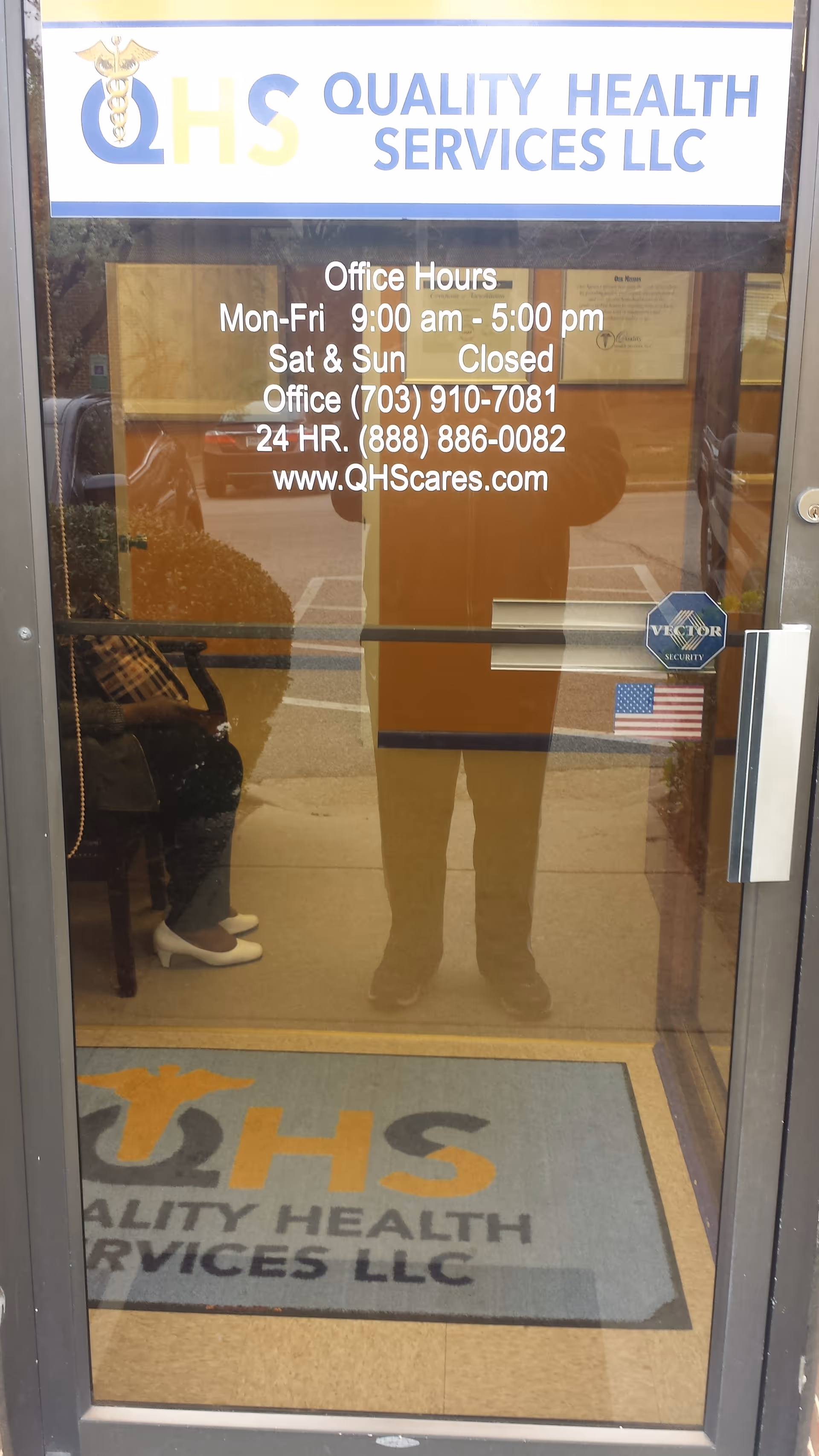 Glass storefront entrance for Quality Health Services LLC with hours posted and a reflected person.