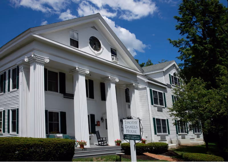 Exterior view of a large white building with classical columns and black shutters under a blue sky with some clouds. A sign in front reads 'Timothy Daniels House, A Skilled Nursing & Rehabilitation Facility'. There are bushes and trees around the building.