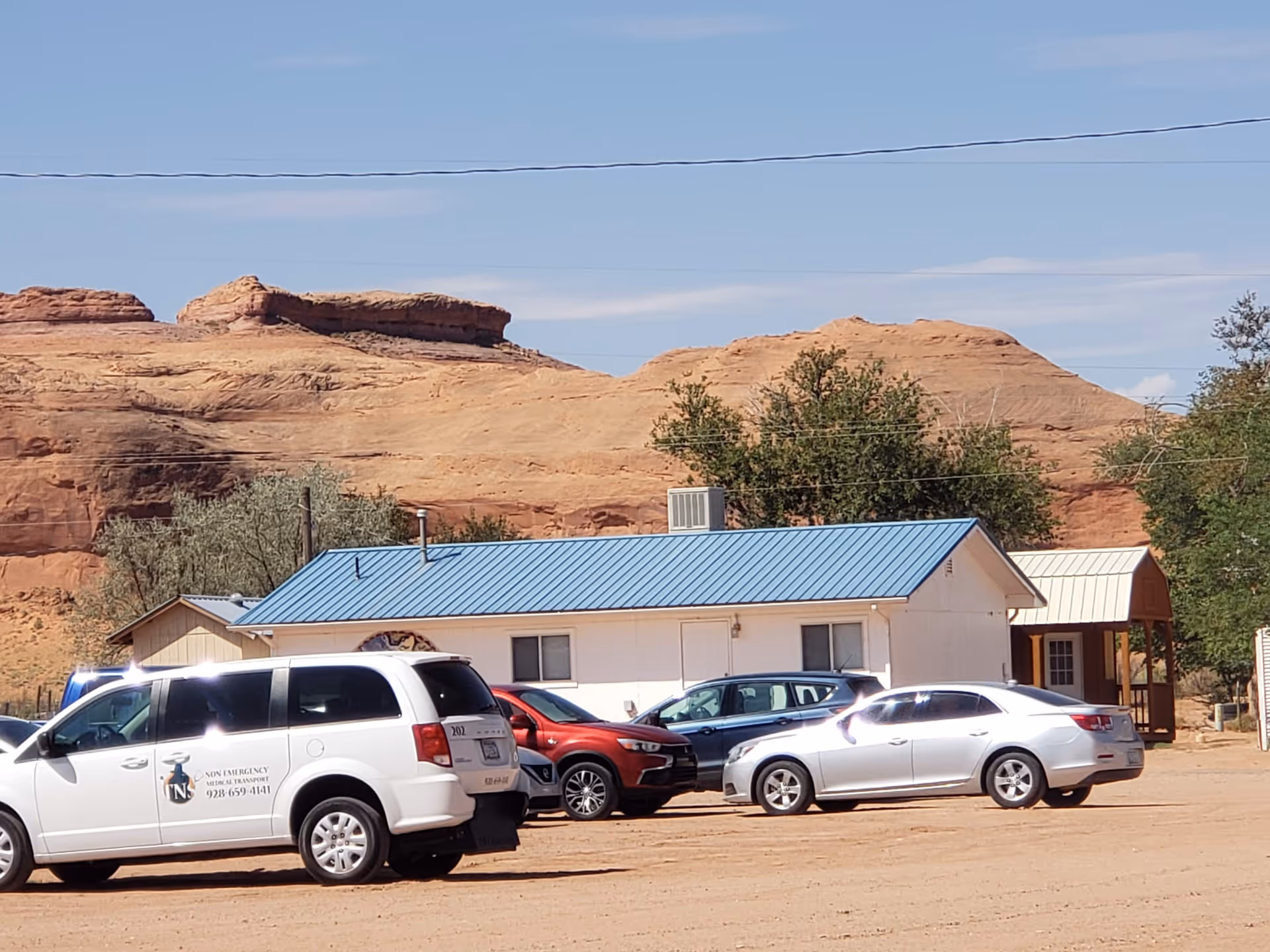 A single-story building with a blue metal roof sits beside desert rock formations and cars.