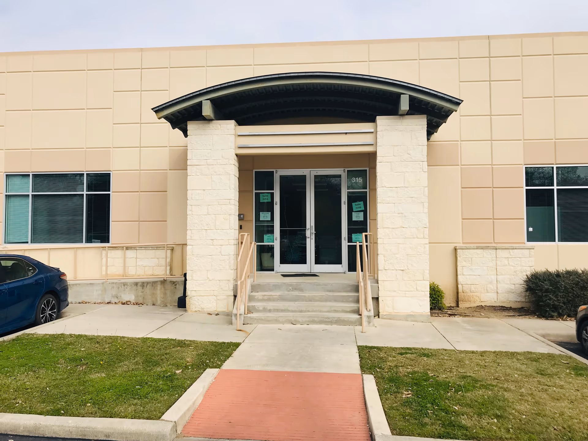 Exterior view of a beige facility entrance with double doors, steps, and ramps.