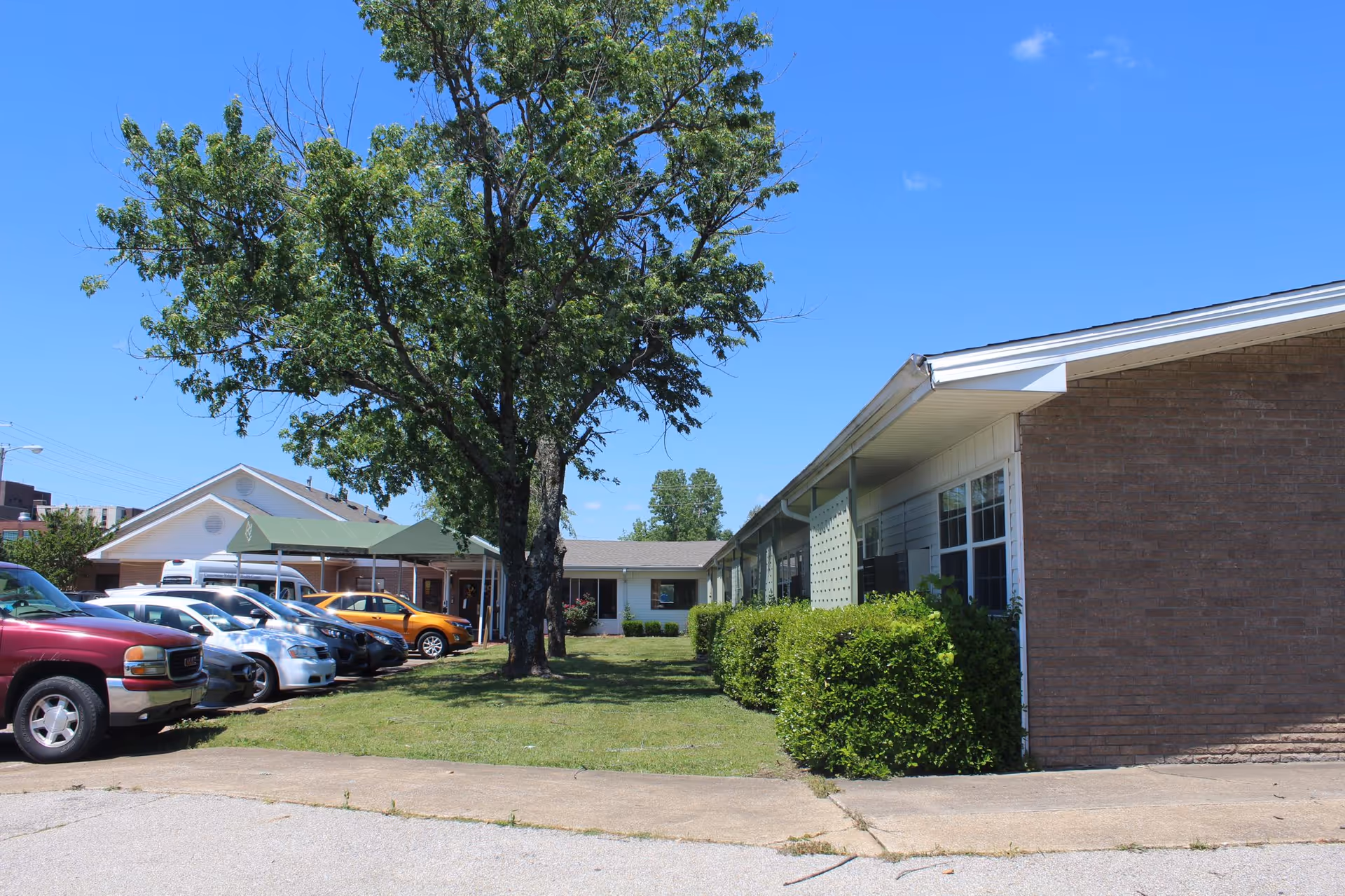 Exterior view of Cornerstone Rehabilitation and Healthcare Center showing a single-story building with a brick and siding facade, a large tree in the grassy area, several parked cars, and a clear blue sky.