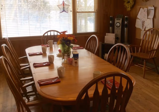 A wooden dining table set for six with red napkins and white mugs, a vase with colorful flowers in the center, and a canister of salt. The room has wooden chairs, large windows with blinds, a star-shaped decoration hanging in the window, and a bulletin board with papers on the wall.
