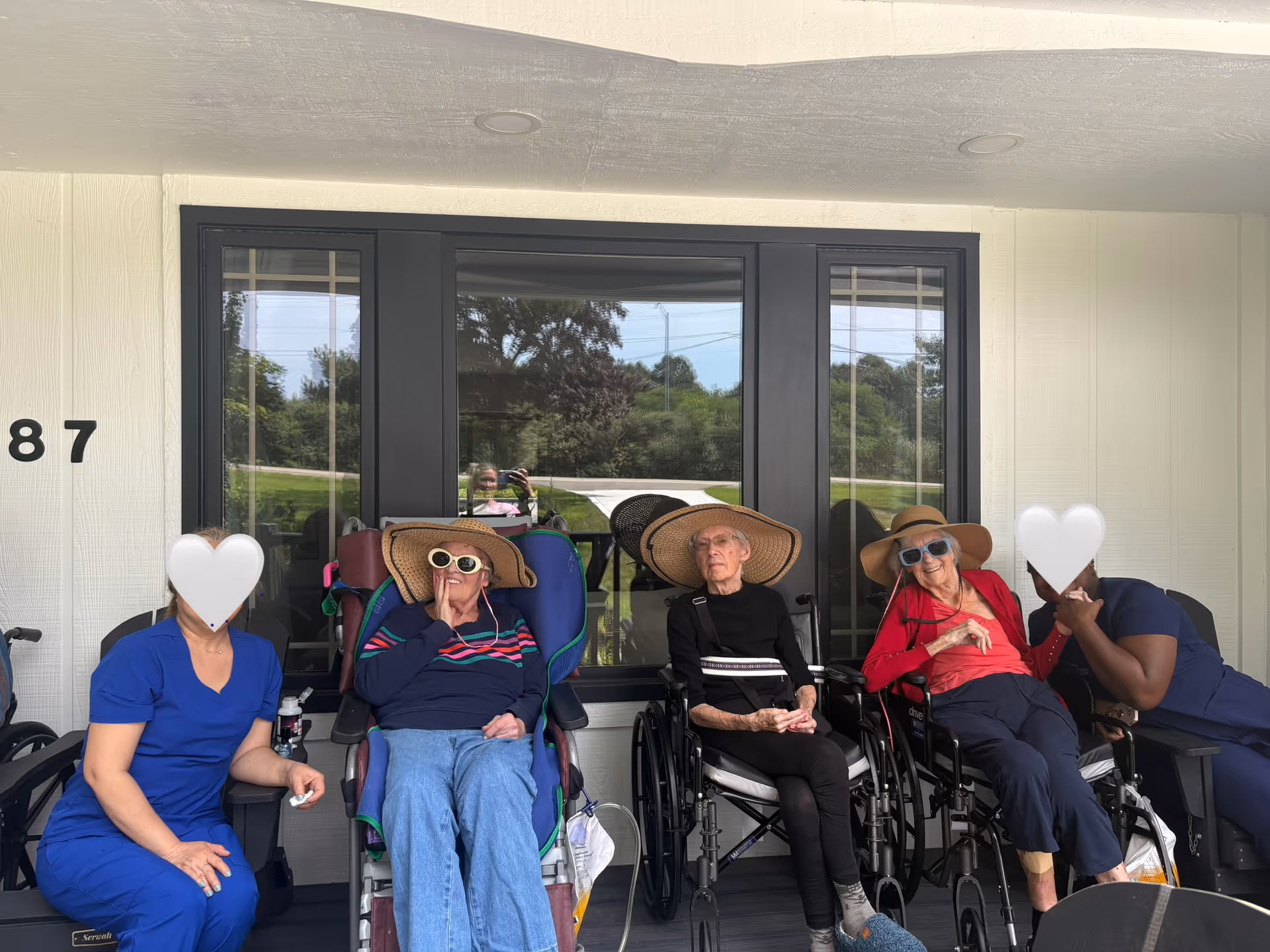 Several seniors in wheelchairs sit on a covered porch outside a building, wearing sun hats.