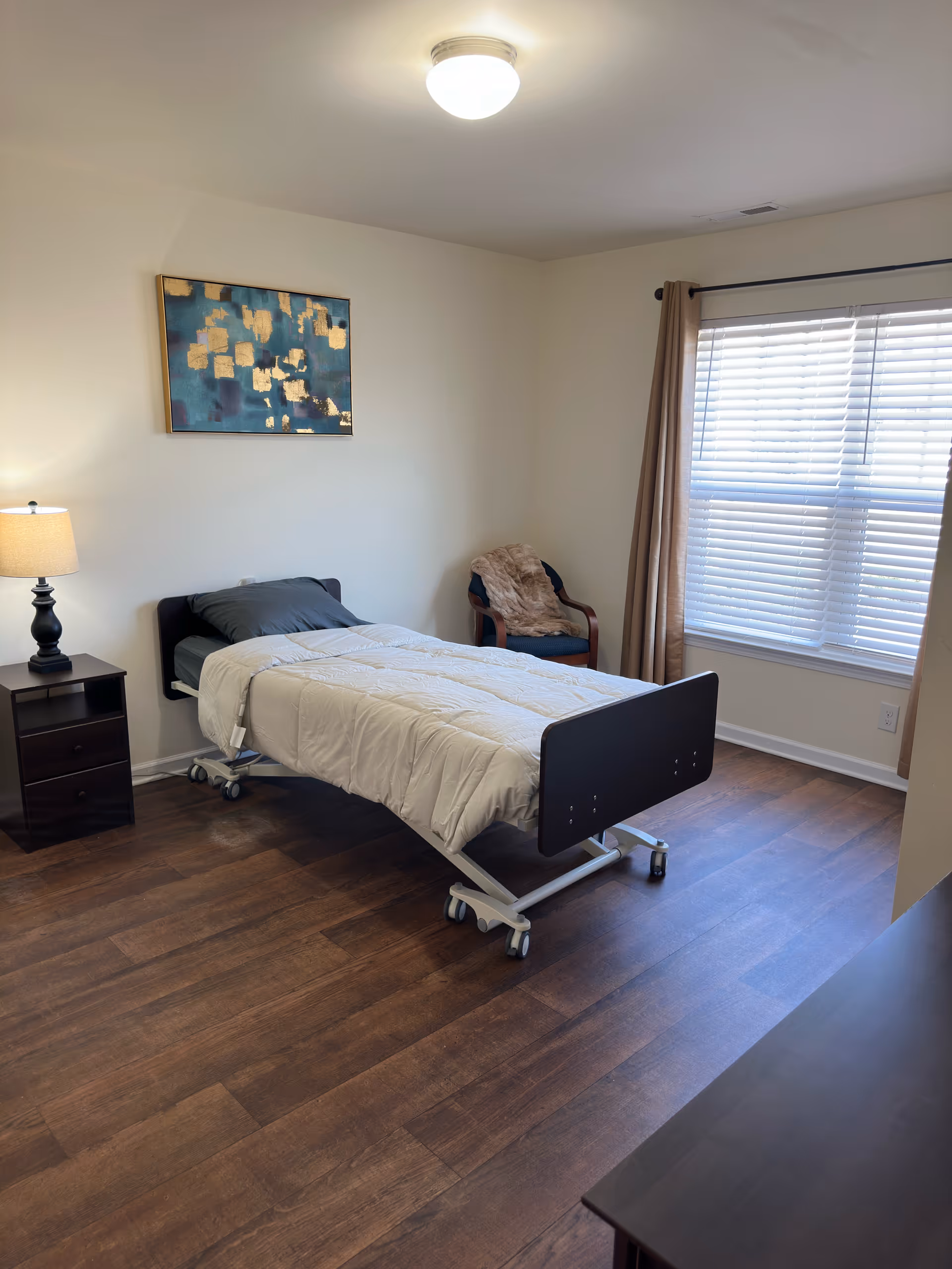 A bedroom in a senior home with an adjustable bed, nightstand, chair, and window blinds.
