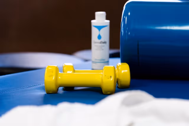 Close-up of two small yellow dumbbells on a blue exercise mat with a blue foam roller and a bottle of lotion or sanitizer in the background.