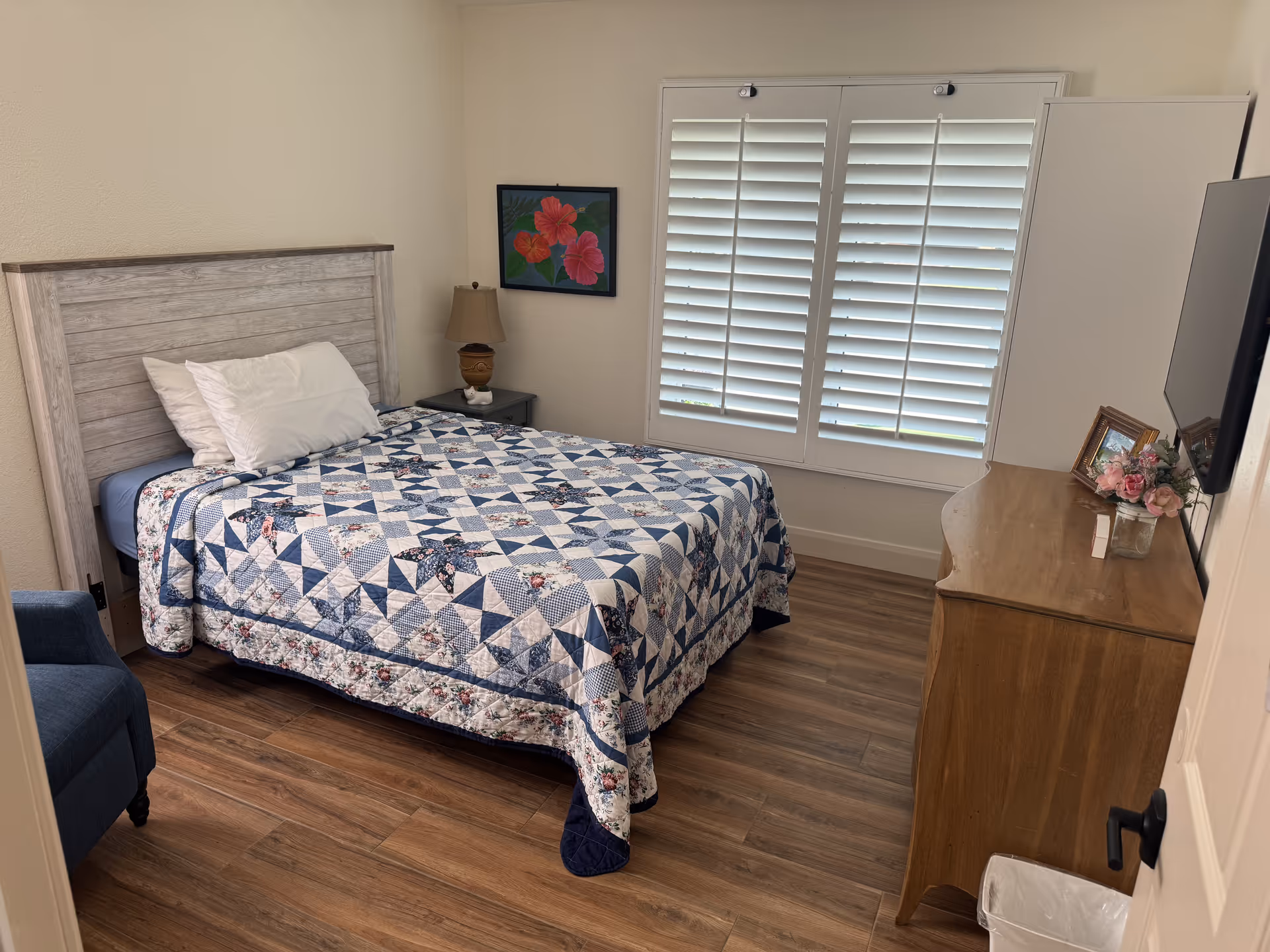 A cozy bedroom with a quilted bed, nightstand, dresser, and window blinds.