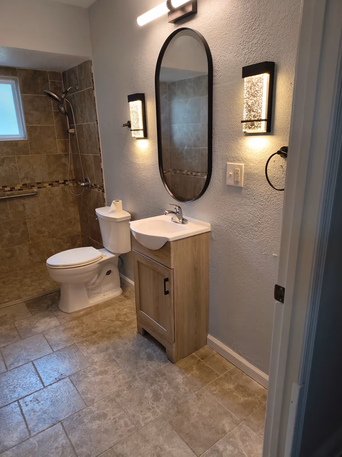 Bathroom with toilet, sink vanity, oval mirror, and walk-in shower in tiled space.
