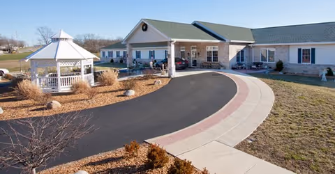 Single-story senior living facility exterior with curved driveway, gazebo, and parked car.