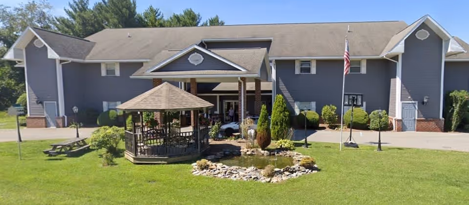 Exterior view of a two-story assisted living facility building with gray siding and a gabled roof. In front of the building is a small landscaped area with a gazebo, a pond surrounded by rocks, shrubs, and an American flag on a flagpole. The building has multiple windows and a covered entrance.