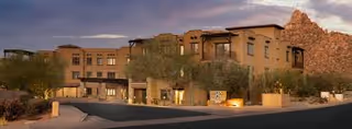 Front exterior of a multi-story Southwestern-style senior living building with desert landscaping and rocky hills at dusk.
