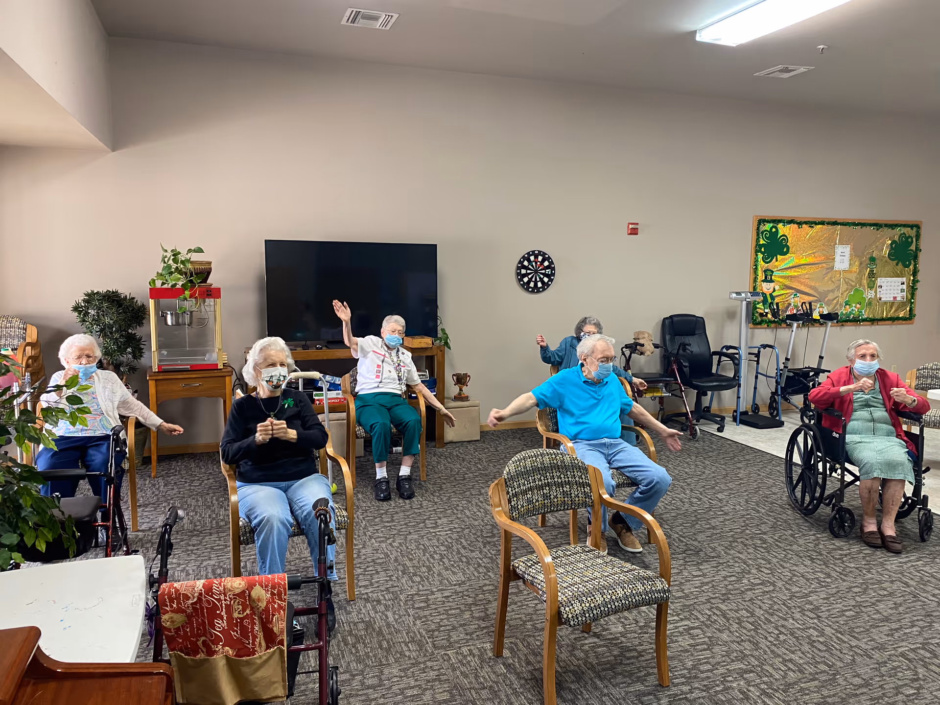 A group of elderly individuals seated in a common room, socially distanced and wearing face masks, participating in a seated exercise or activity. The room has a large TV, a dartboard on the wall, a popcorn machine, and a bulletin board decorated with shamrocks.