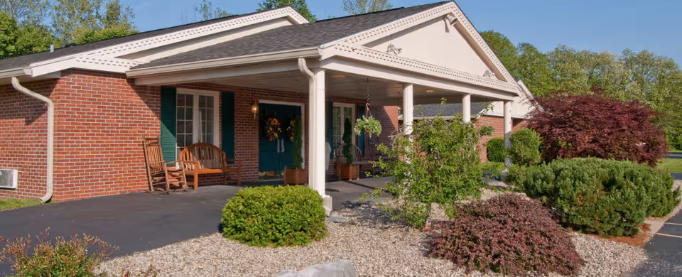 Exterior view of a single-story brick building with a covered entrance supported by white columns. There are two wooden rocking chairs and a bench near the entrance door, which is decorated with a wreath. The surrounding area features well-maintained landscaping with various bushes, shrubs, and trees under a clear blue sky.