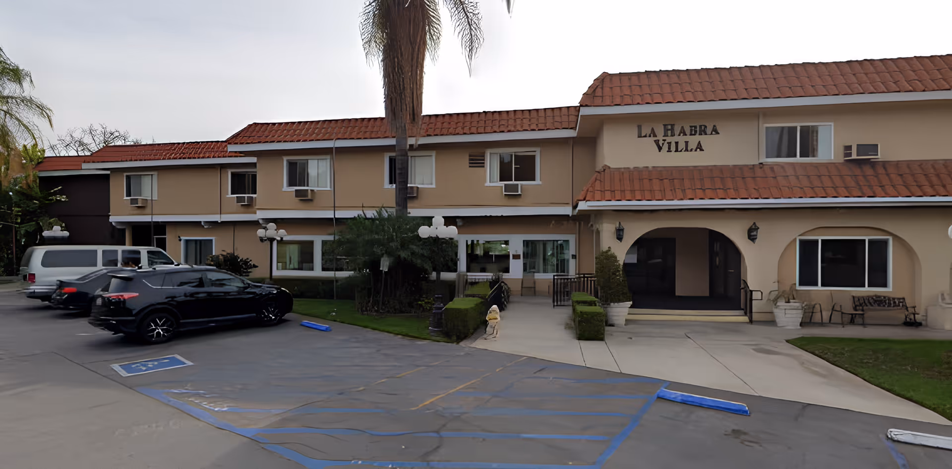 Front exterior of a two-story beige senior living building labeled "La Habra Villa" with parked cars, a palm tree, and handicapped parking spaces.