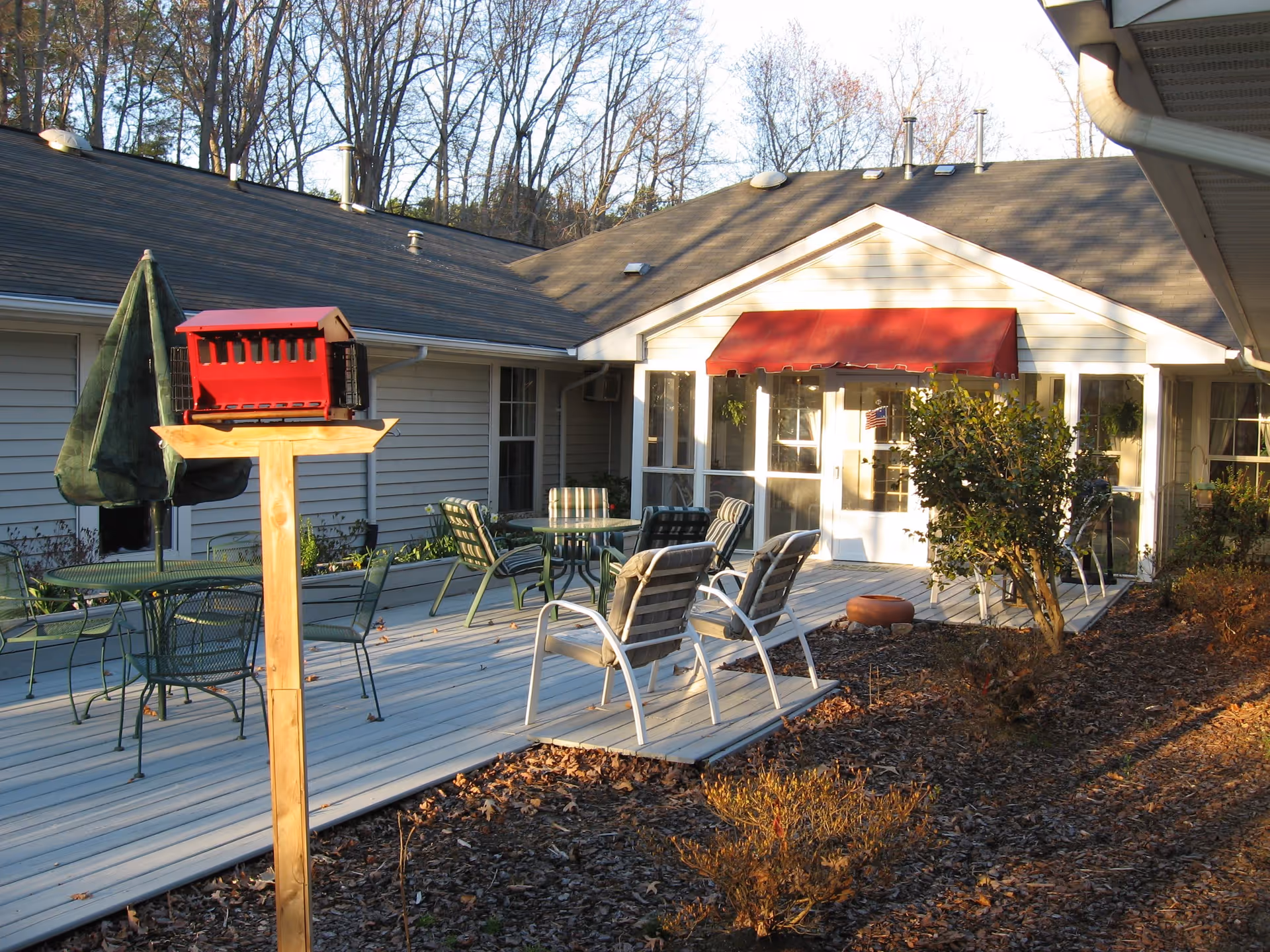 Outdoor patio area at Bayberry Retirement Inn featuring several metal tables and chairs, a red bird feeder mounted on a wooden post, a closed green umbrella, and a building with a red awning over a glass door entrance. Trees without leaves are visible in the background.