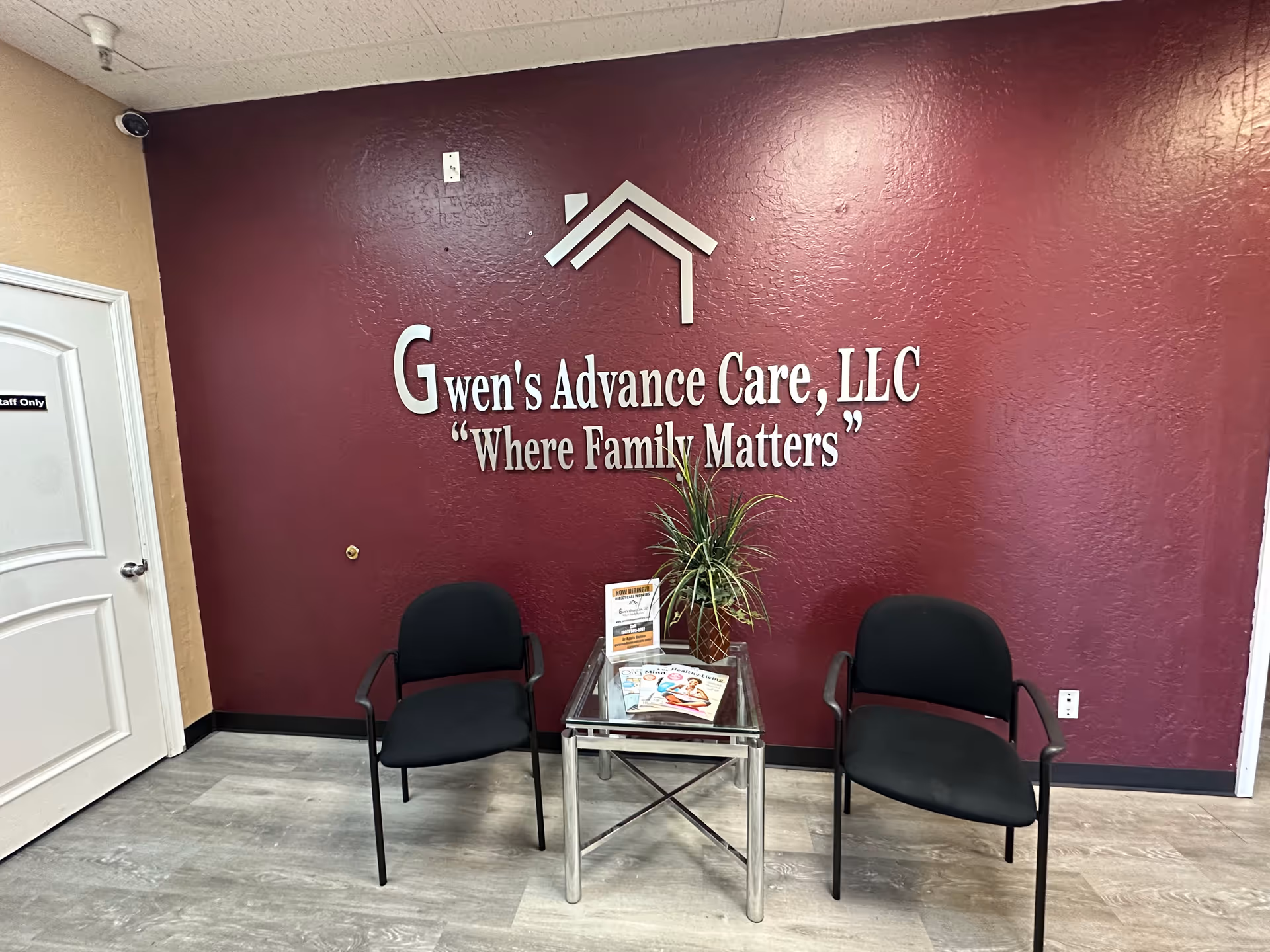 Interior lobby with maroon wall displaying Gwen's Advance Care logo, two chairs, and a glass table.