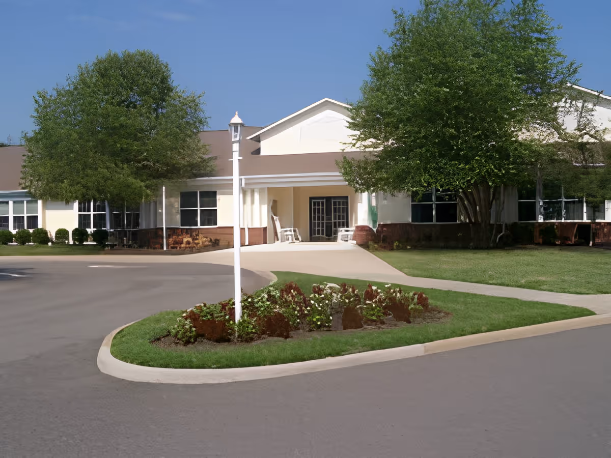 Front exterior view of a single-story building with a driveway, landscaped flower bed, and large green trees under a clear blue sky.