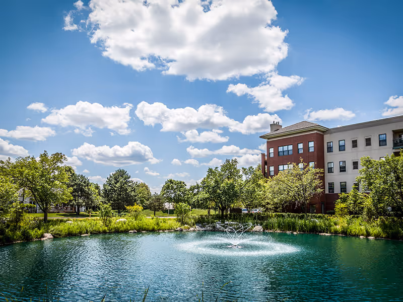 A scenic outdoor view of a pond with a water fountain in the center, surrounded by green trees and shrubs. In the background, there is a multi-story building with red brick and beige exterior walls under a blue sky with scattered white clouds.