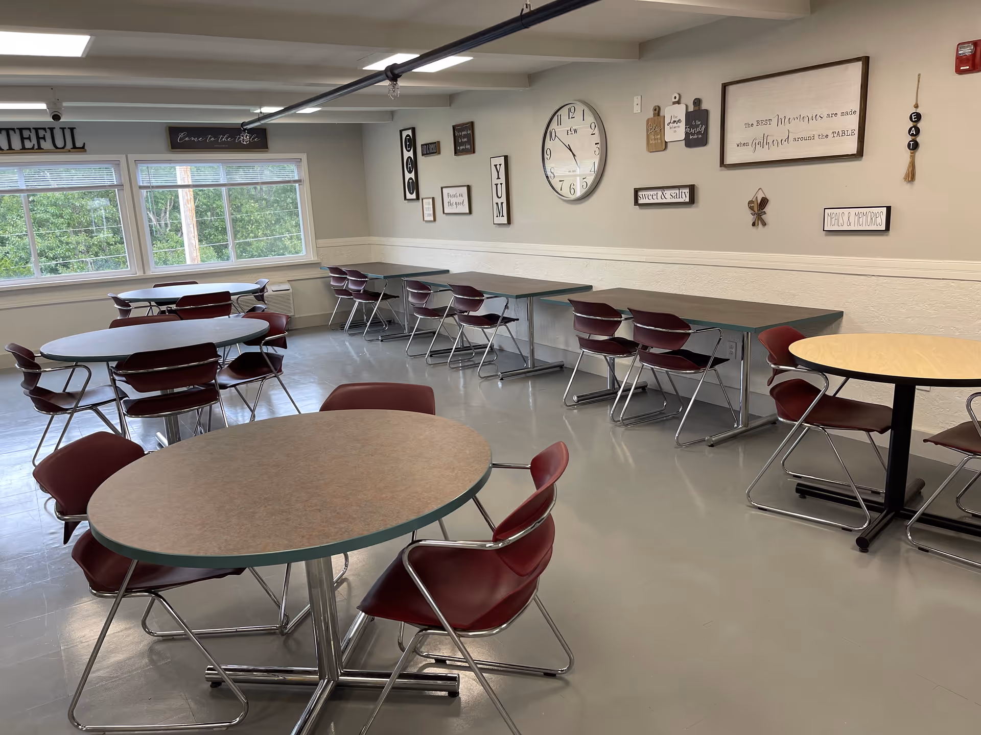 Bright communal dining room with round tables, maroon chairs, and wall decor.