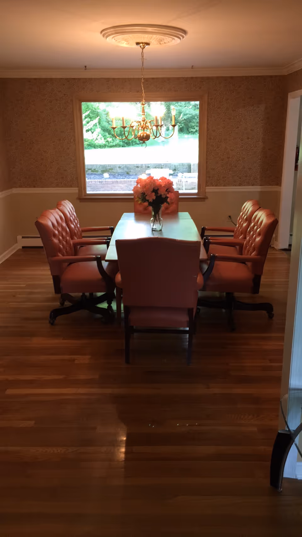 A dining room with a wooden table surrounded by six cushioned chairs with armrests. A vase with pink flowers is placed in the center of the table. A chandelier hangs above the table, and there is a large window in the background showing greenery outside. The room has wooden flooring and wallpapered walls with a chair rail.