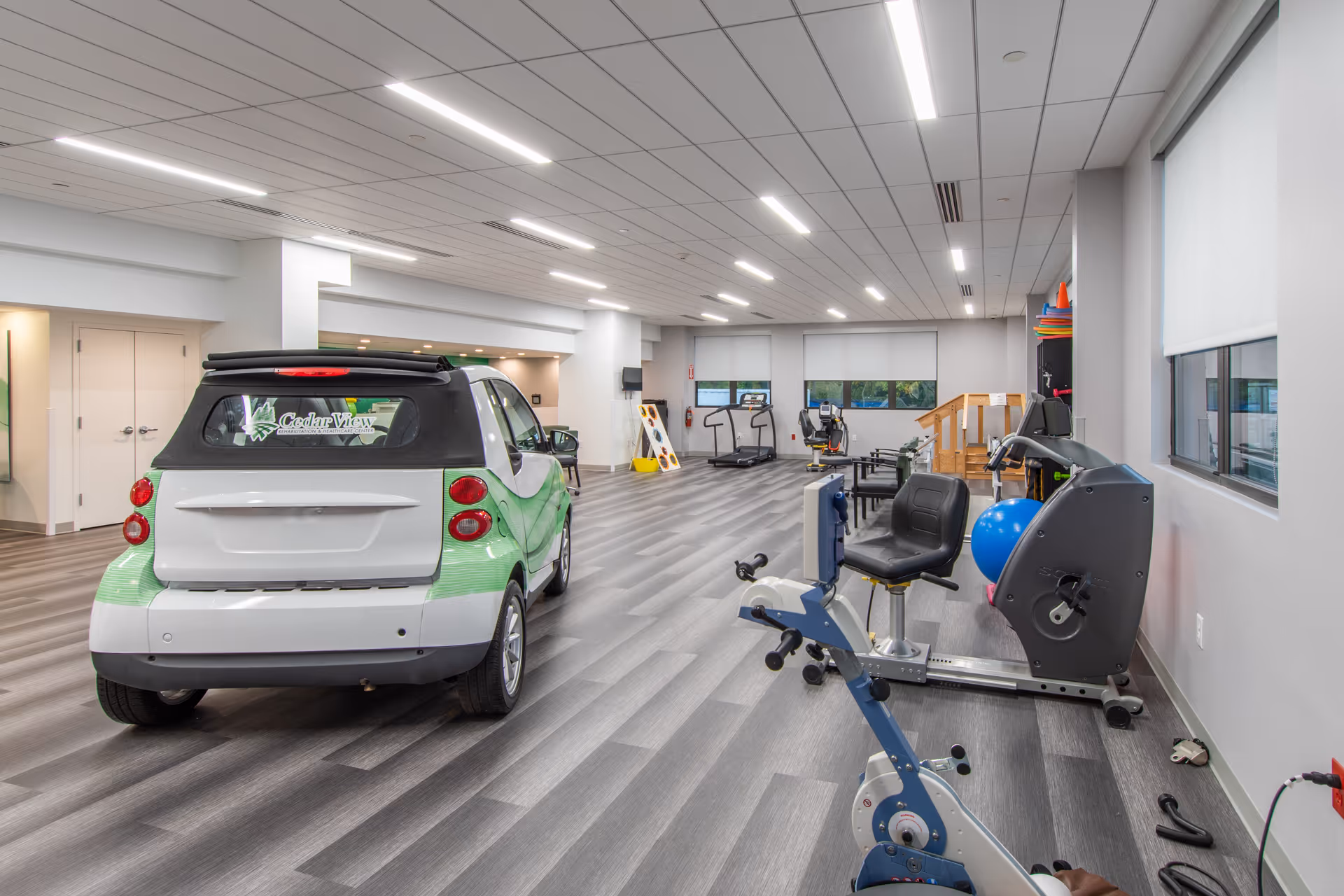 Spacious rehabilitation gym interior featuring exercise machines, therapy equipment, and a small branded car parked on a wood-look floor.