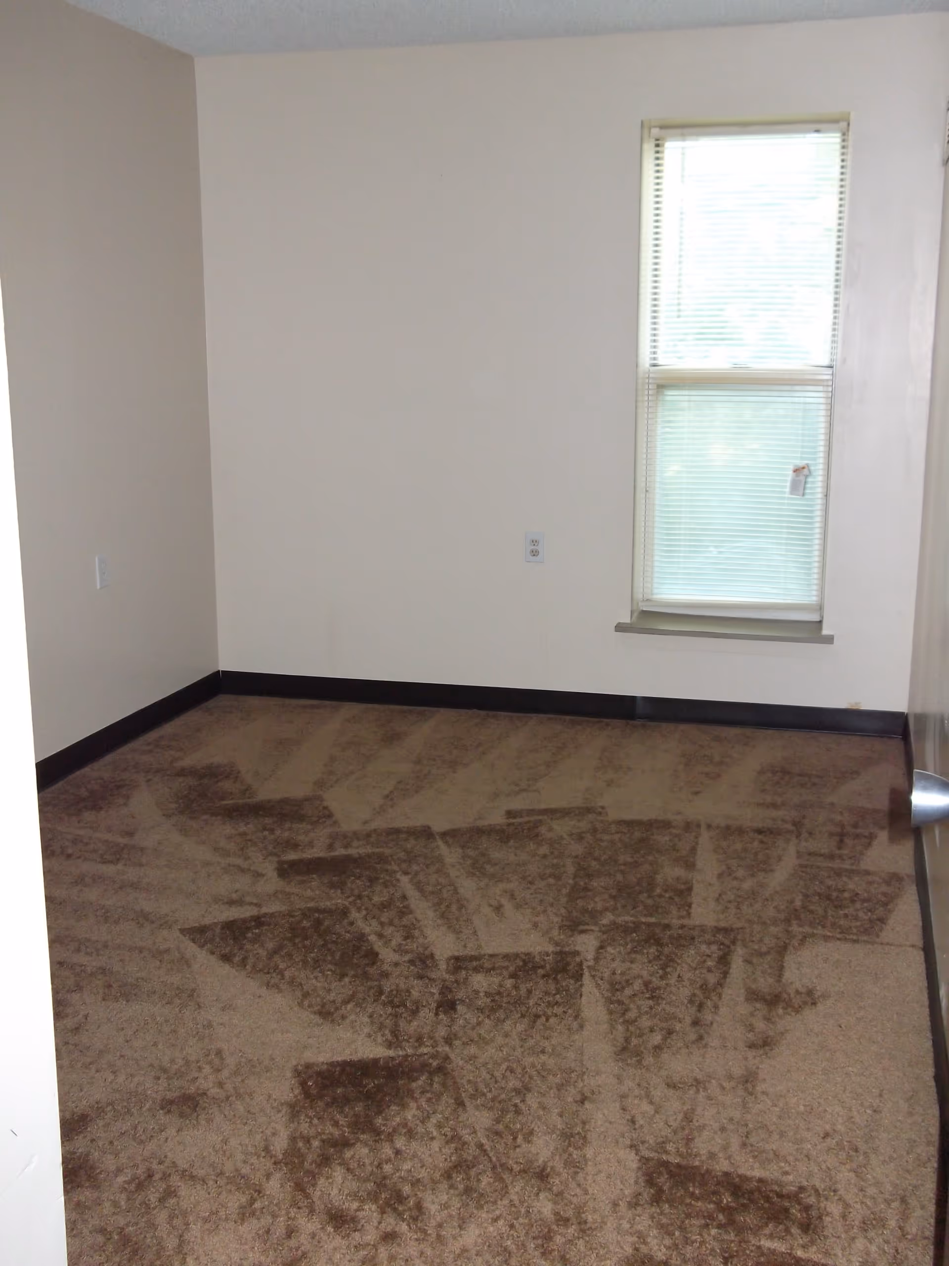 Empty small room with beige walls, a single window with closed blinds, and patterned brown carpet.