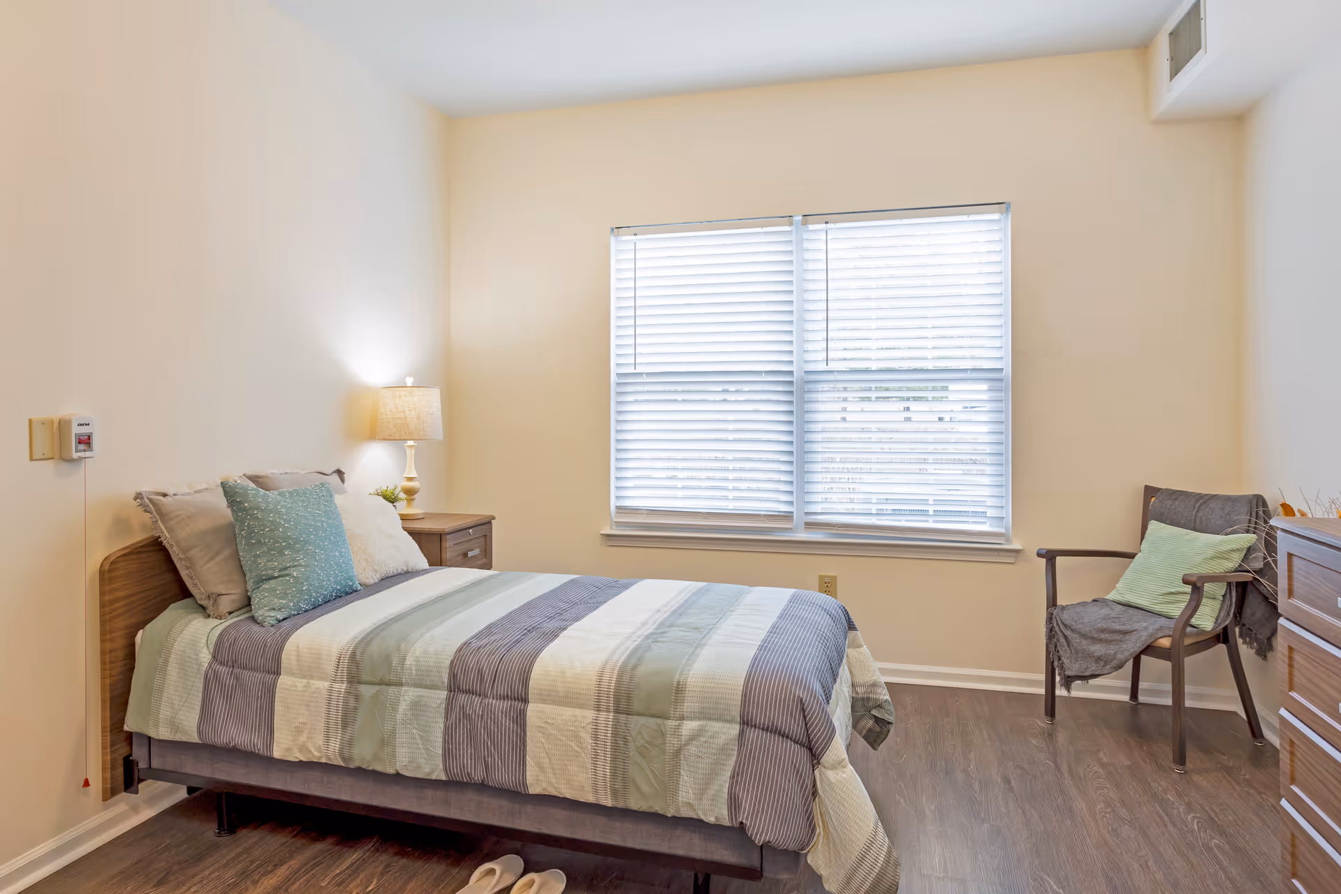 A neatly made bedroom with a bed, nightstand, lamp, window blinds, and chair.
