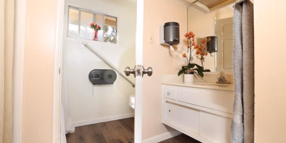 Accessible bathroom with a white vanity and sink, wall-mounted dispensers, grab bar, and a toilet visible through an open door.