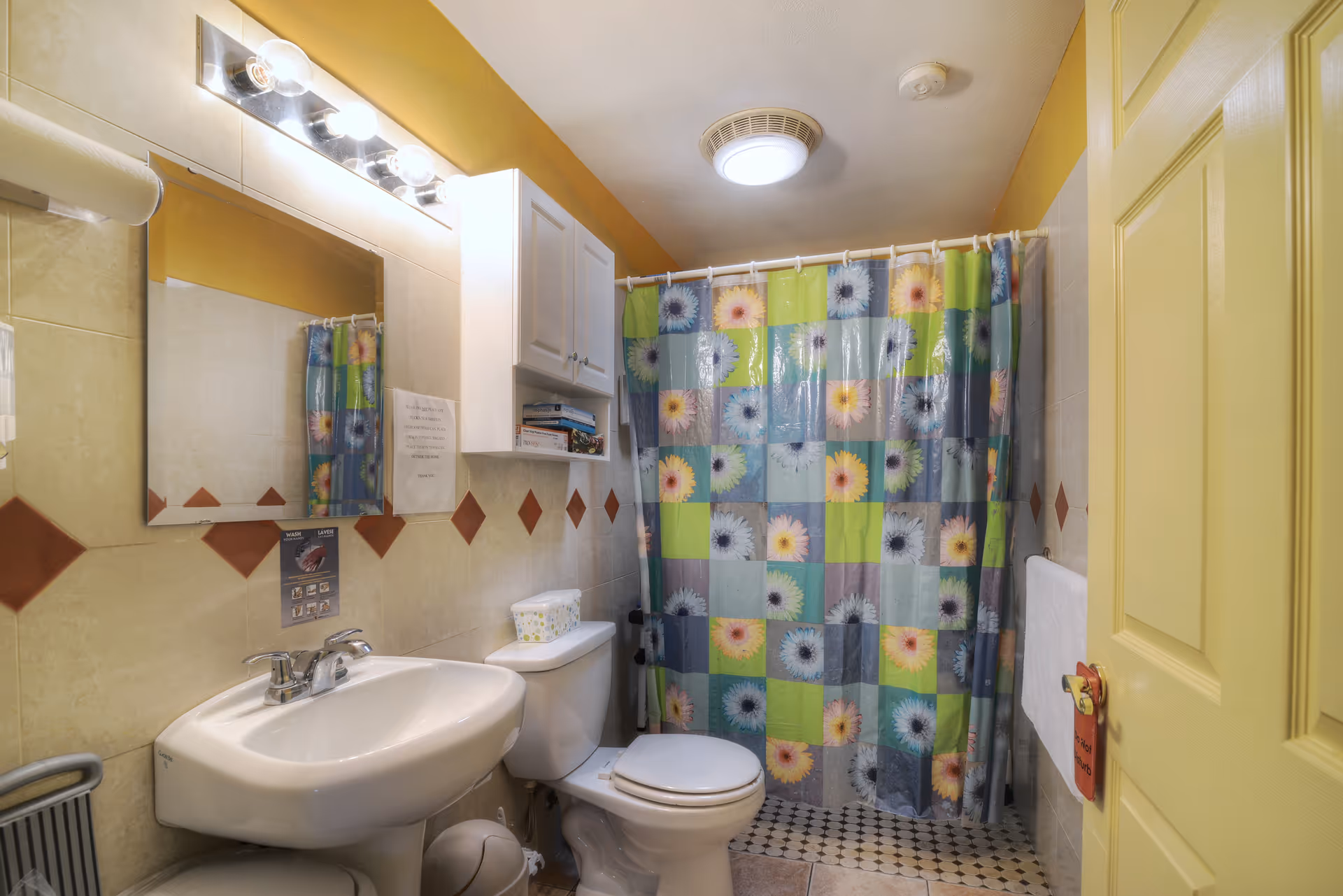 A small bathroom with a white sink, toilet, and a colorful shower curtain featuring a floral patchwork design. Above the sink is a mirror with lights, and a white cabinet is mounted on the wall. The walls have beige tiles with a row of diamond-shaped brown tiles. A yellow door is partially open, and a towel hangs on a rack beside it.