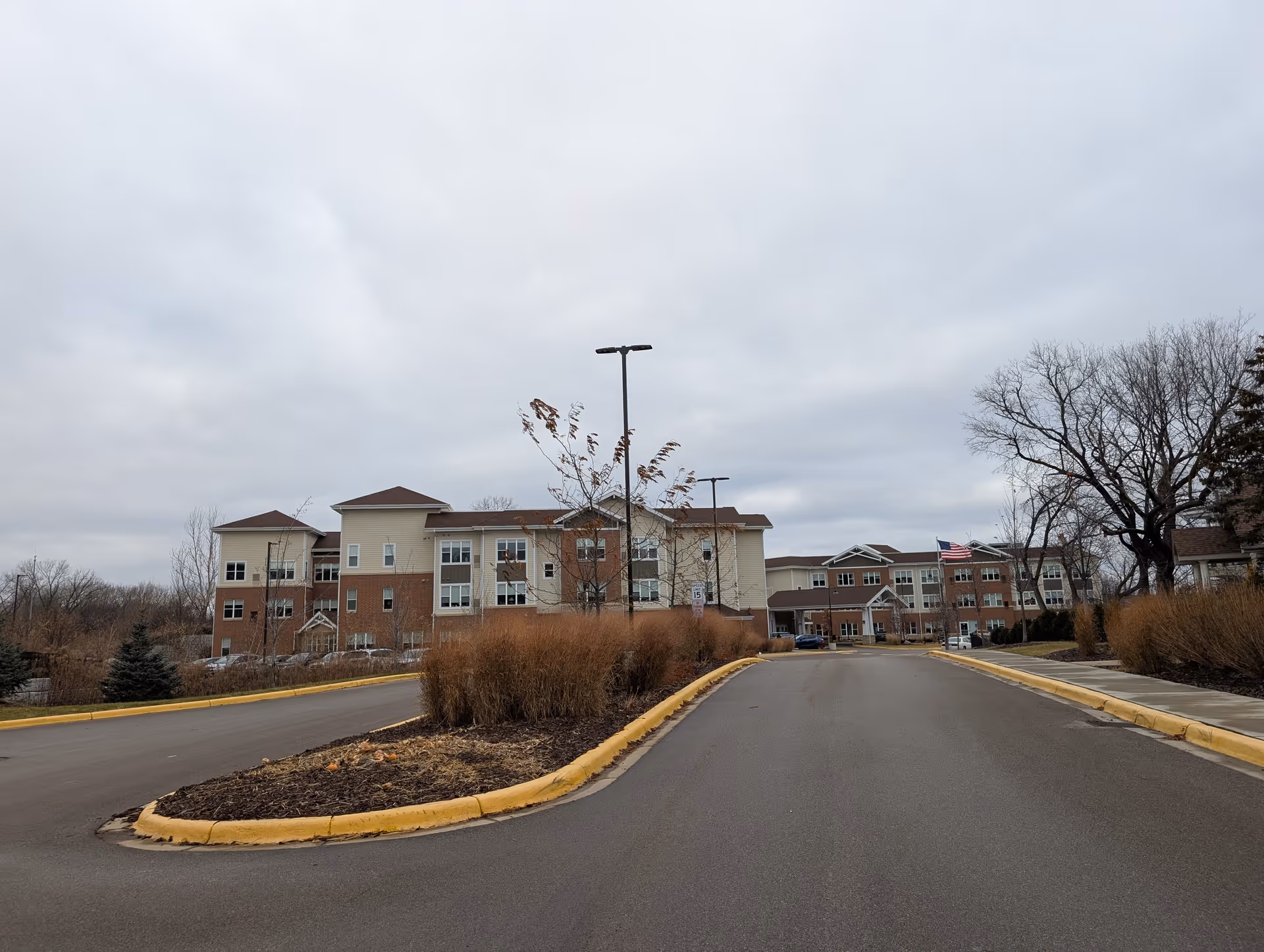 Front entrance and driveway of a multi-story senior living building with landscaping under an overcast sky.