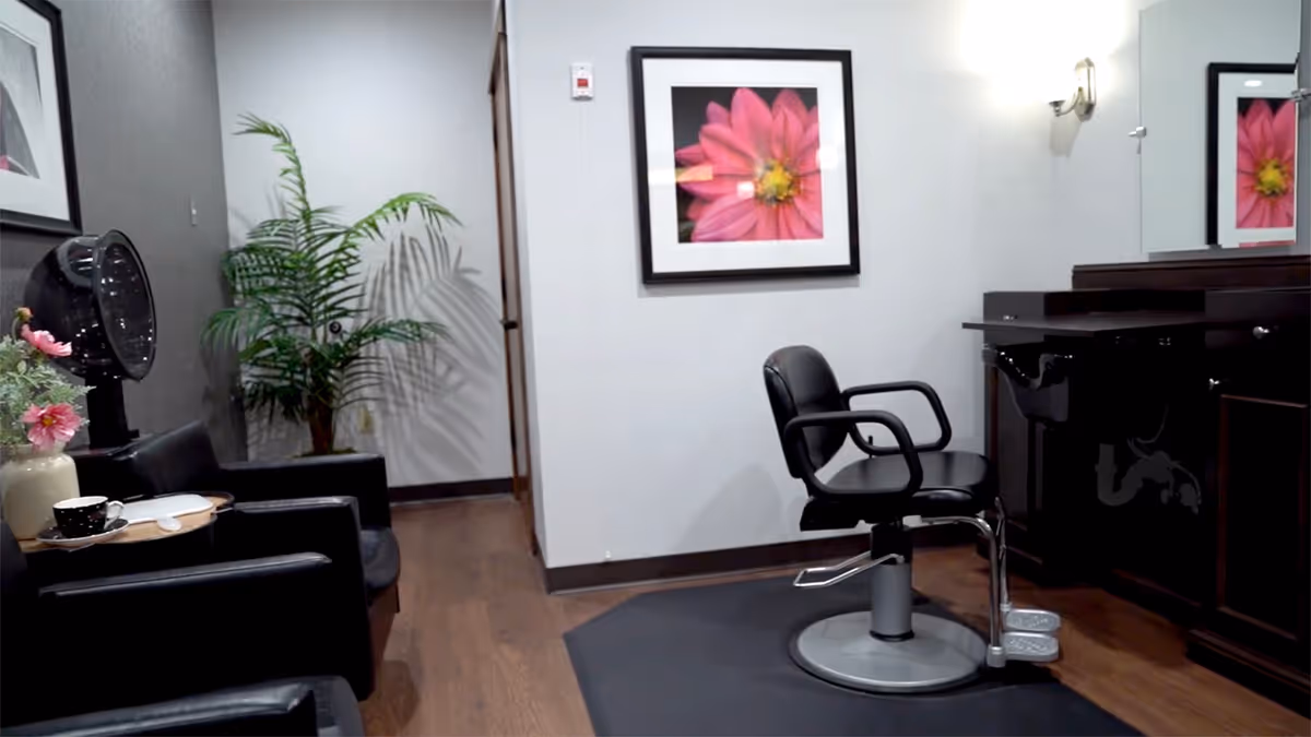 Interior salon area with black styling chair, mirror, plant, and framed pink flower art.