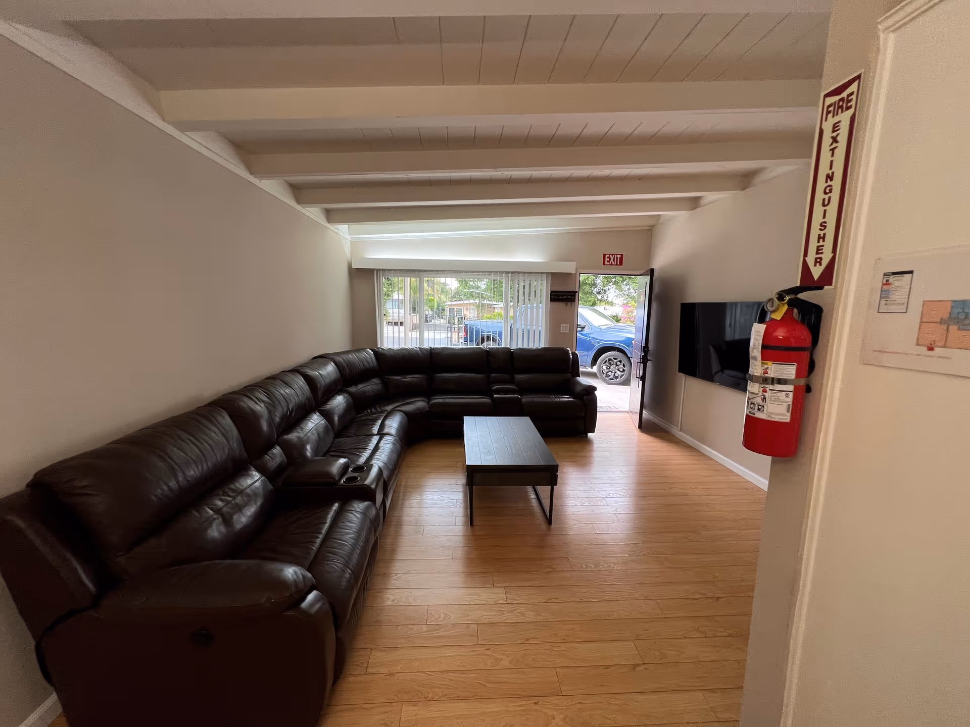 Communal living room with a large brown leather sectional sofa, coffee table, wall-mounted TV and an open exit door.
