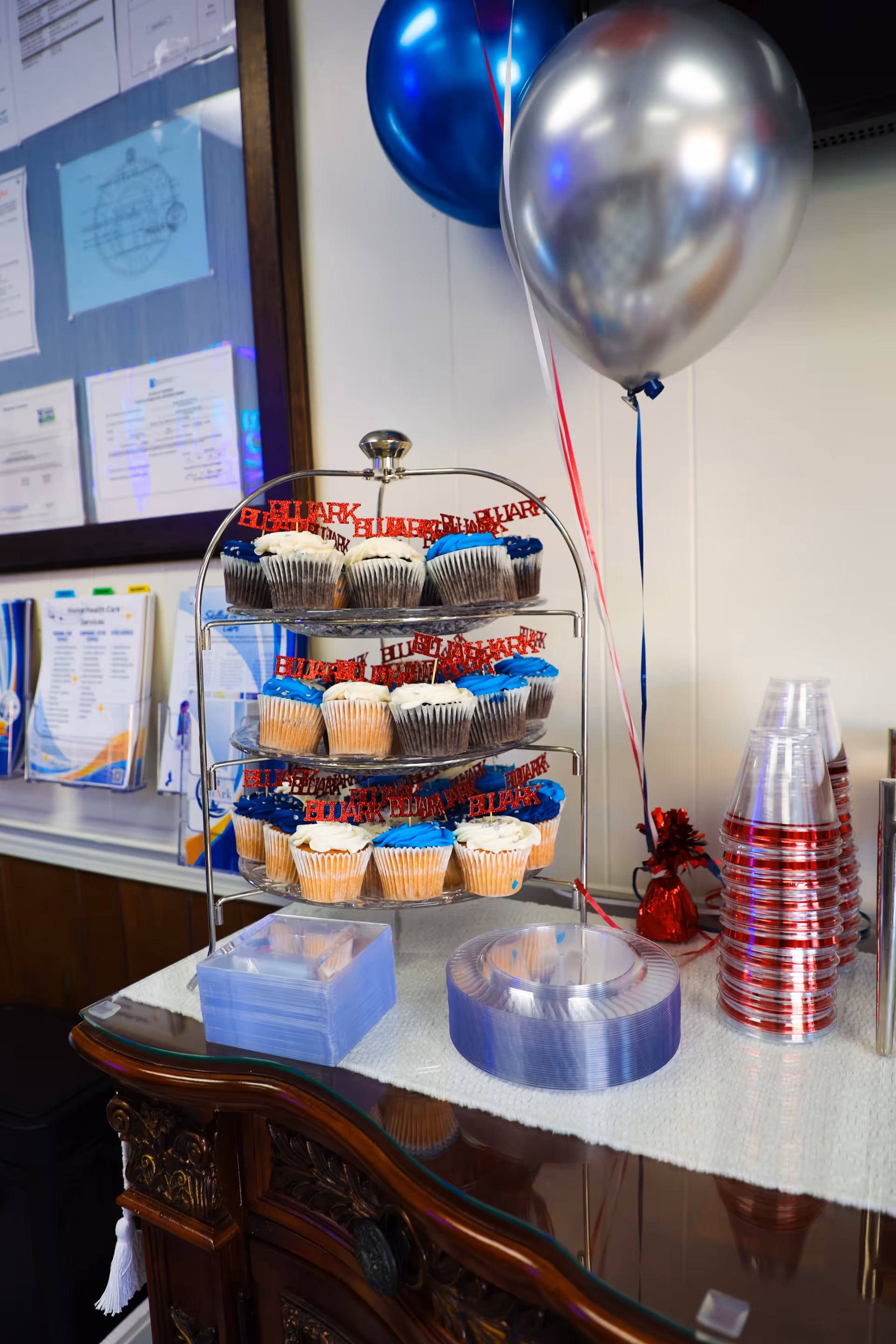 Assorted cupcakes displayed on a metal stand with balloons beside a cup stack indoors