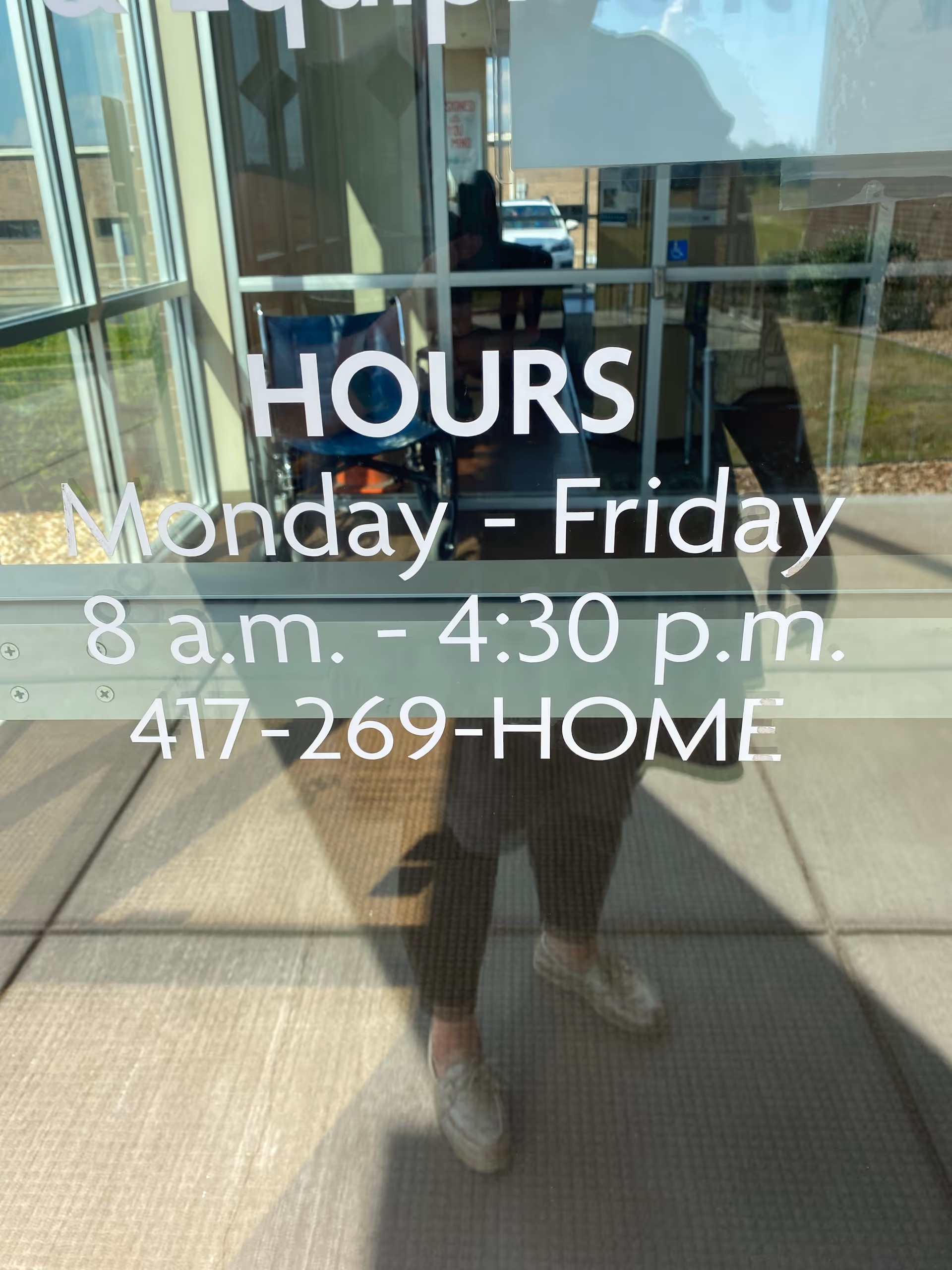 Glass front door displays hours sign with a reflection of a person outside.