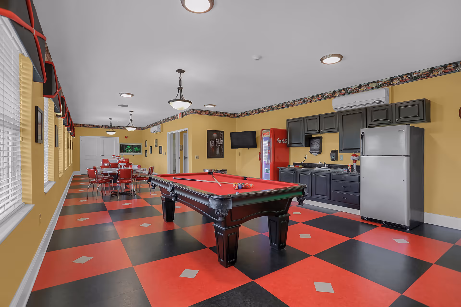 A recreational room with a red and black checkered floor, yellow walls, and multiple windows with red valances. The room features a pool table with a red felt top in the center, a kitchenette area with dark cabinets, a stainless steel refrigerator, and a red Coca-Cola vending machine. There are also several tables and chairs arranged along the left side of the room, and ceiling lights providing illumination.