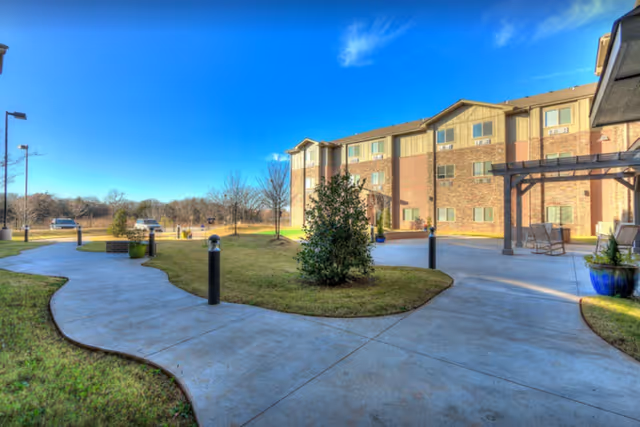 Curved concrete walkways and landscaped courtyard in front of a three-story brick senior living building under a blue sky.
