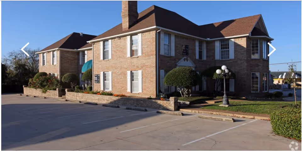 Brick senior living facility front with manicured shrubs and a parking lot.