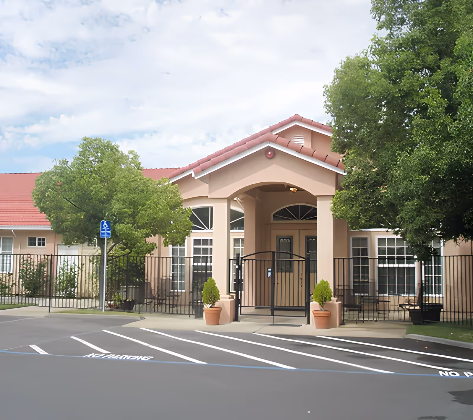 Front entrance of a single-story senior living building with a covered portico, gated entry, potted plants, and marked parking spaces.