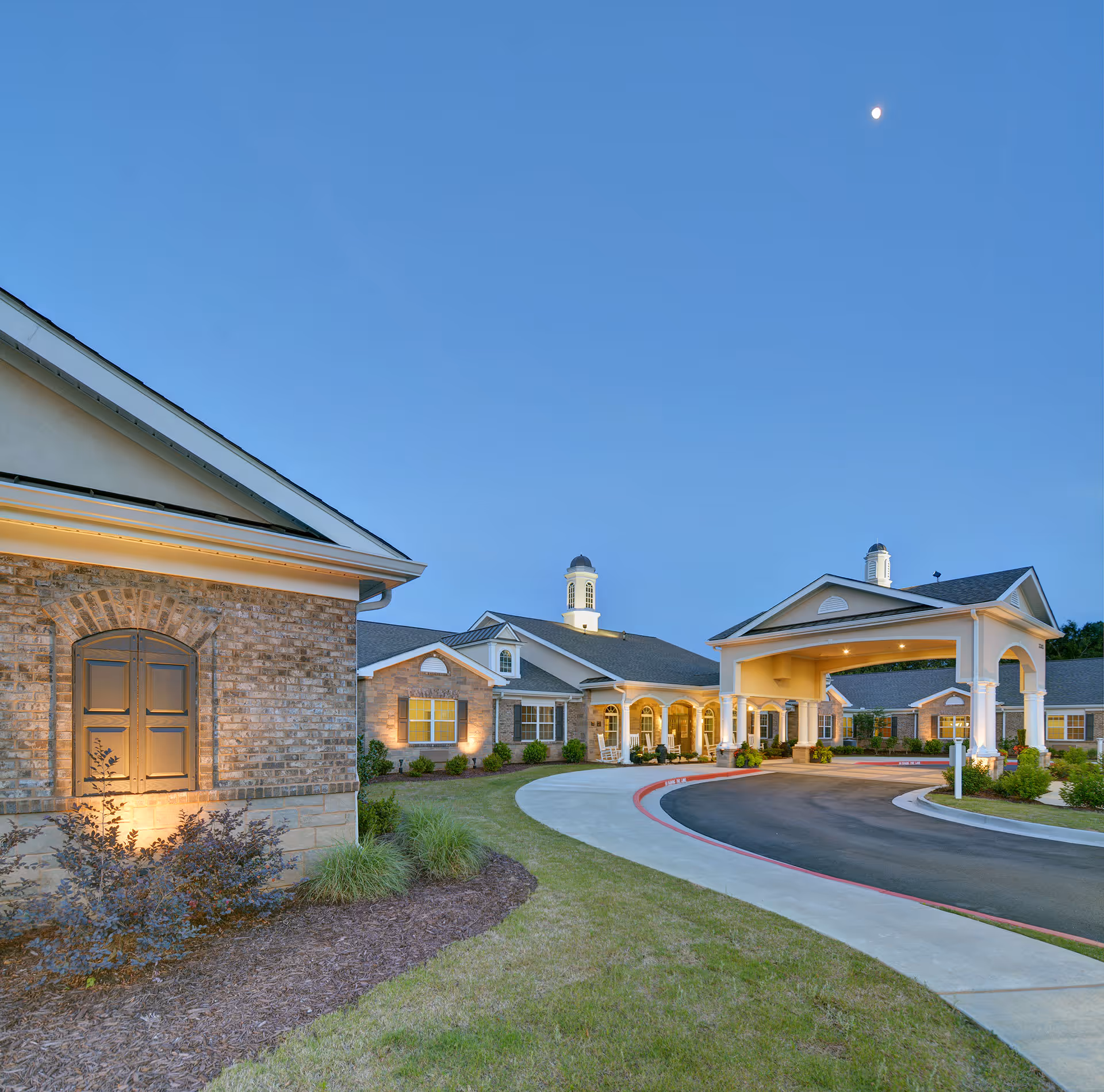 Exterior view of Benton House of Woodstock senior living facility at dusk, showing a curved driveway leading to a covered entrance with columns, well-maintained landscaping, and a clear sky with a visible moon.