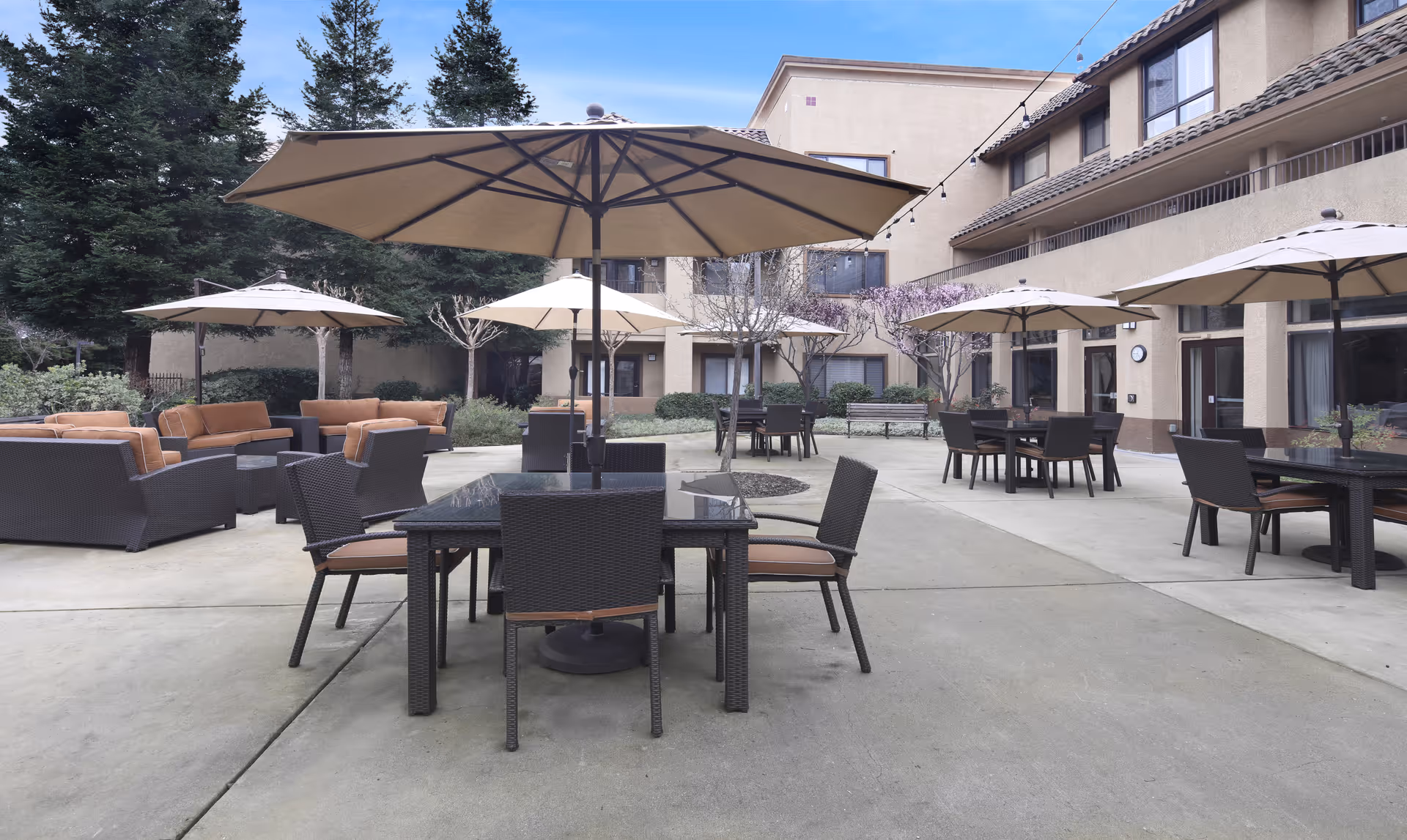 Outdoor patio area at Ivy Park at Roseville with multiple tables and chairs under large beige umbrellas, surrounded by a beige building and trees in the background.